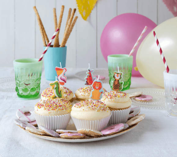 Image of a kids party table set up.  In the forefront is a plate of 5 cupcakes with animal candles in them. 