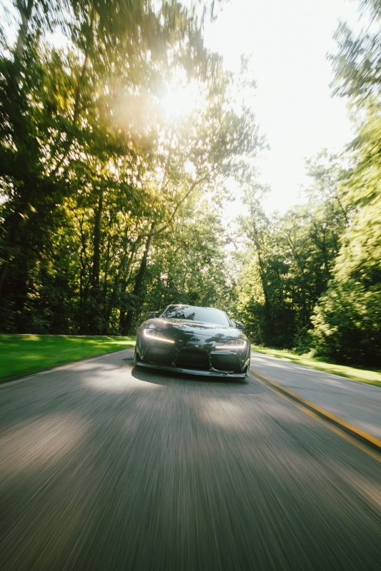 A black sports car driving toward the camera at high speed, on a country road with the sun behind the treeline