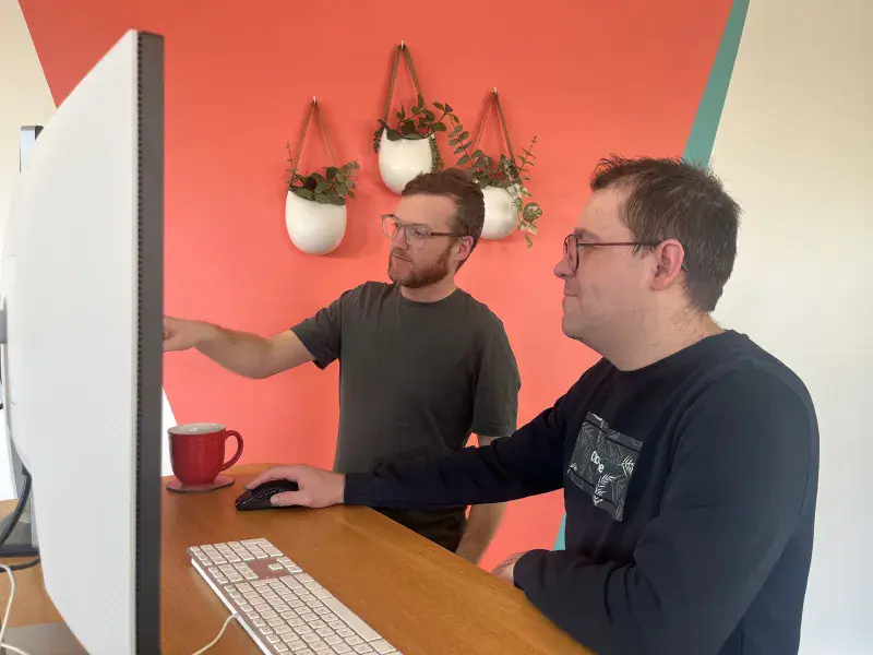 Two web developers stand at a desk and solve a problem together on the screen