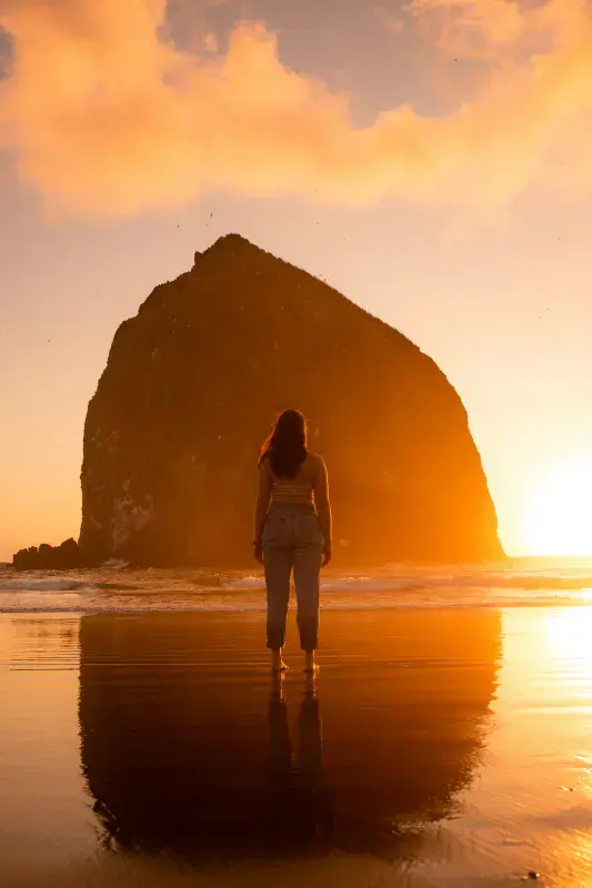 Woman stands on a beach at sunrise looking out, the sun peeking from behind a tall island