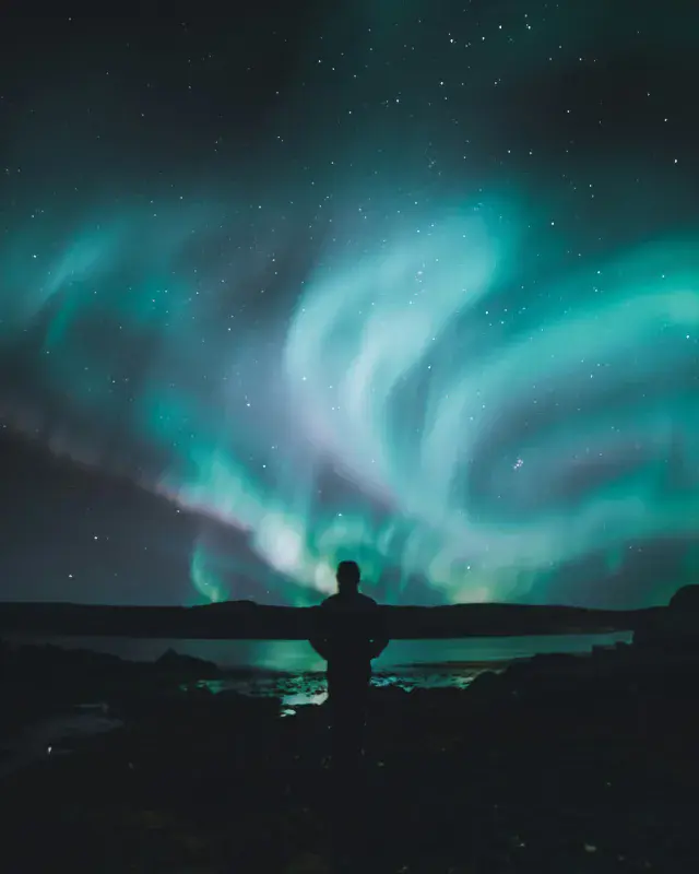 Silhouette of a man standing in front of a lake at night, swirling aurorae lighting up the sky