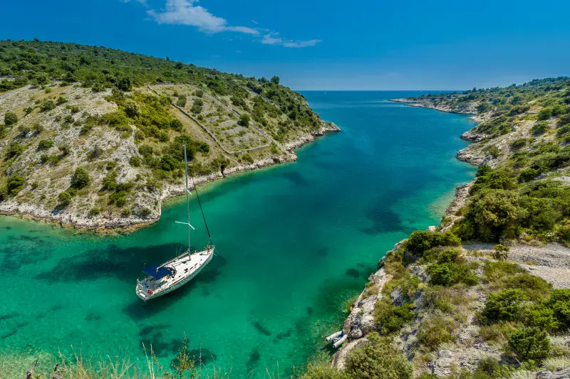 Sailboat anchored in a turquoise bay surrounded by rocky, green hills under a clear blue sky.