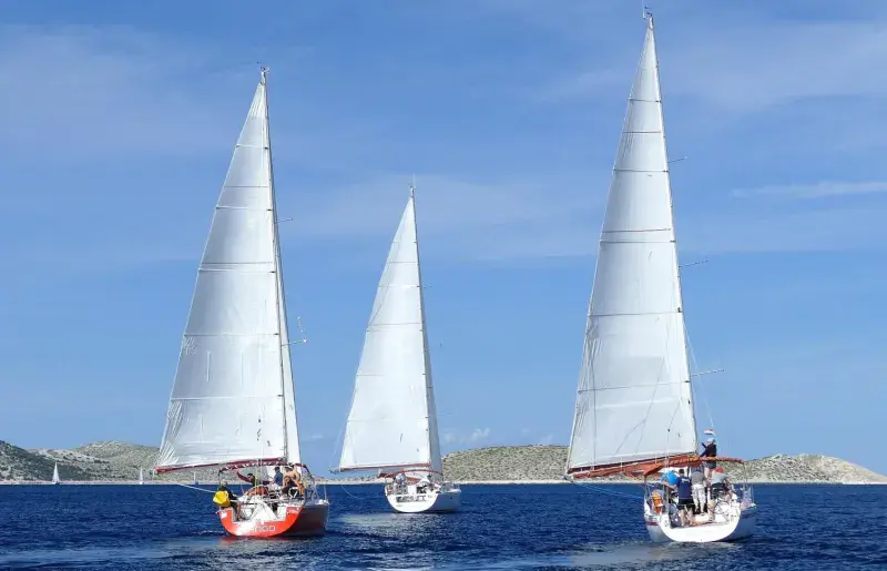  Three sailboats with full white sails are sailing on a choppy blue sea, with a rocky coastline visible in the background under a clear blue sky.