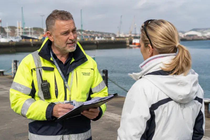Harbour master in a high-visibility jacket and ID badge talking with a female yacht captain in a harbour.