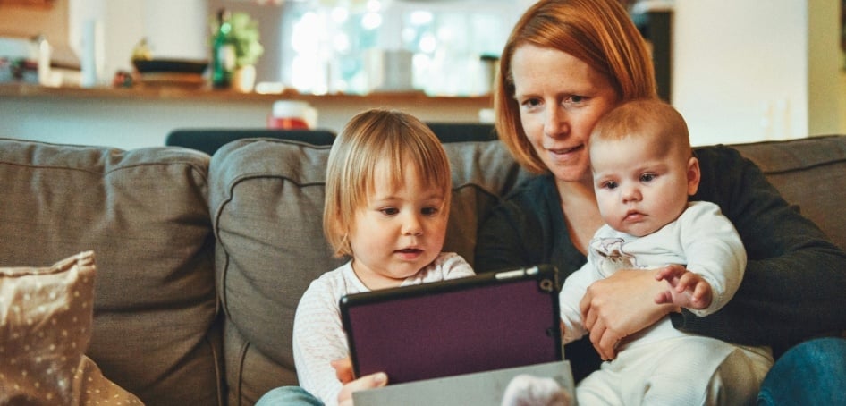 Mum sits at home on the sofa with her toddler and baby, looking at an iPad together