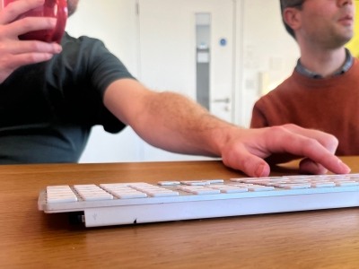 Web developer typing on a keyboard with one hand, drinking coffee with the other, a colleague sat with him