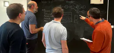 A group of software engineers gather around a blackboard wall, one is pointing at part of the diagram