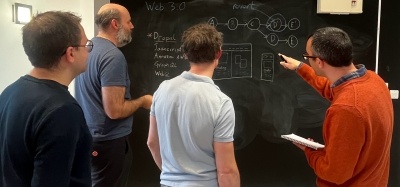 A group of software engineers gather around a blackboard wall, one is pointing at part of the diagram