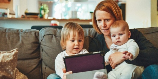 Mum sits at home on the sofa with her toddler and baby, looking at an iPad together