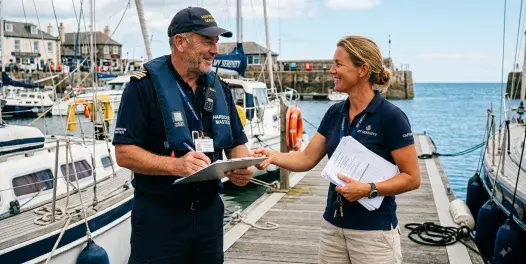 A male harbour master in official uniform, lifejacket, and cap writes on a clipboard while meeting with a female yacht captain holding paperwork on a wooden pier, with docked boats and harbour buildings in the background.